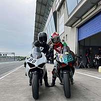 Zwei Fahrer in Rennanzügen neben ihren Motorrädern auf der Rennstrecke.