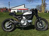 Ein Ducati Scrambler Motorrad auf einer Wiese mit Hintergrundgebäuden.