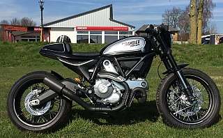 Ein Ducati Scrambler Motorrad auf einer Wiese mit Hintergrundgebäuden.